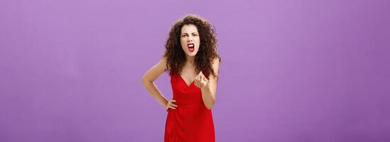 Woman in fury fighting with husband over money making italian gesture with fingers shouting and yelling from fury and anger standing displeased and pissed over purple wall in red elegant dress photo