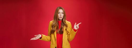 Frustrated and confused young cute female with ginger hair in yellow coat shrugging as pointing questioned left waiting explanation as asking question clueless and puzzled over red background photo