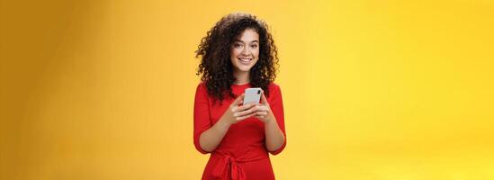 Girl messaging friend to tell all hot rumors holding mobile phone in hands smiling broadly and excited at camera as using smartphone browsing in net or edditing in app over yellow background photo
