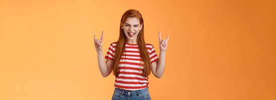 Daring cool excited redhead sassy woman having fun enjoying awesome rock-n-roll concert smiling thrilled enthusiastic grin, squinting sly show heavy-metal signs, stand orange background photo