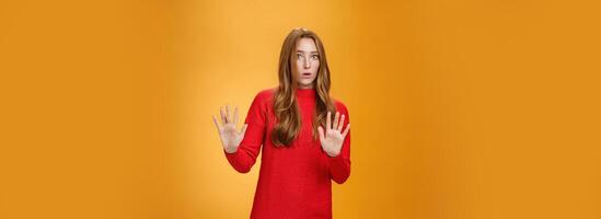 Intense panicking and freaked out girl with red hair asking slow down, raising hands near chest in stop and not gesture open mouth surpised and shocked, dismaying strange offer over orange background photo