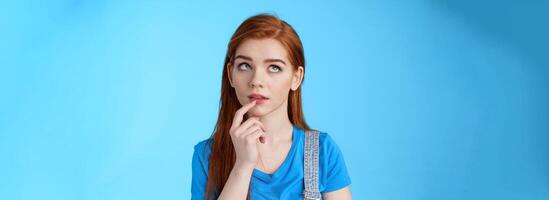 Close-up curious dreamy creative redhead female working project, student thinking essay, touch lip intrigued, look up thoughtful, pondering choice, make assumptions, stand blue background photo