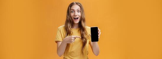 Girl surprised with awesome smartphone features. Portrait of delighted and amazed cute european slender woman with wavy natural hairstyle showing cellphone pointing at gadget screen at app photo