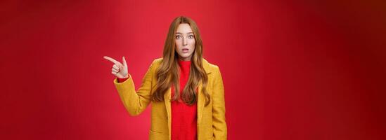 Impressed and questioned amazed redhead woman remain speechless from amazement pointing right open mouth in stupor, asking question about confusing shocking promotion over red wall photo