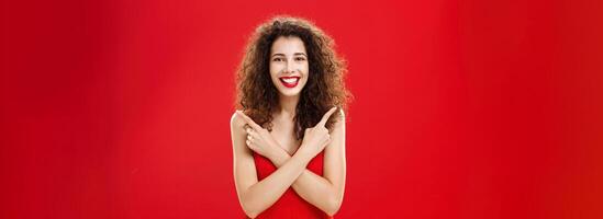 Woman satisfied with any decision feeling carefree and happy standing in elegant red evening dress with curly hairstyle crossing hands on chest pointing left and right delighted with choices photo