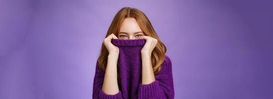 Girl awaits winter with happy grin being ready warming up with cozy sweater pulling collar on face and smiling with eyes at camera standing positive and cute against purple background photo