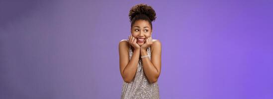 Amused silly cute black girl in silver dress anticipating exciting party dreamy gaze up touch cheeks smiling broadly optimistic waiting dream come true standing pleased touched blue background photo