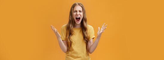 Studio shot of young woman during argument losing temper standing pressured and pissed yelling out loud from hate and anger gesturing with raised palms making furious face posing against orange wall photo