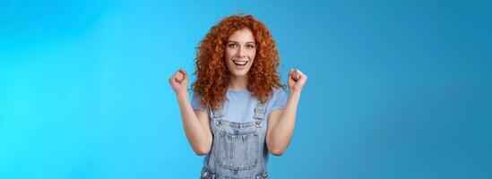 Hooray successful day. Cheerful chrismastic redhead girl summer dungarees clench fists joyfully smiling broadly determined achieve success, triumphing winning game standing upbeat blue background photo
