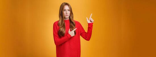 Intense suspicious and doubtful european ginger girl in warm sweater frowning hesitant and confused pointing at upper left corner asking question, looking with disbelief over orange background photo