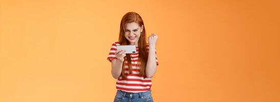 Pleased lucky redhead girl gamer redhead, fist pump satisfied, say yes excited stare display, hold smartphone horizontal, pass hard level playing awesome game use application, achieve victory photo