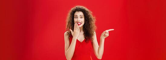 Troubled cute and tender silly woman with curly hairstyle in red stylish evening dress saying oops smiling with guilty look covering mouth with palm while pointing left over studio background photo
