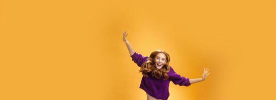 Expressive and enthusiastic carefree friendly joyful redhead woman in purple sweater and pants jumping joyfully with raised hands smiling yelling from happiness and joy against orange background photo