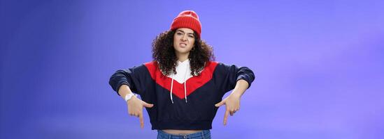 Snobbish and displeased stylish and cool street girl with curly hair in beanie and sweatshirt pointing down grimacing as feeling unsatisfied and disappointed posing against blue background photo