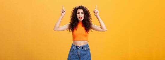 Indoor shot of displeased female client with curly stylish hairstyle red lipstick in cool cropped top pointing up demanding explanation with confused and dissatisfied expression over orange background photo