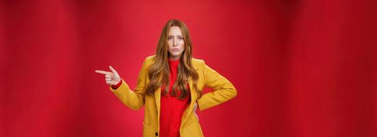 Explain yourself now. Portrait of upset gloomy and sad cute redhead girl waiting explanations asking question, pouting offended and pointing right with finger looking unhappy at camera over red wall photo