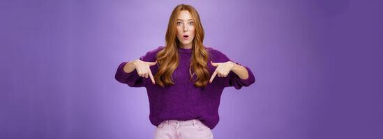 Hold on what is that. Portrait of questioned and thrilled impressed cute redhead female with freckels open mouth from interest as asking question about awesome product, pointing down photo