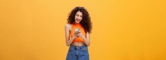 Girl spending time in internet texting friend messages via smartphone laughing while looking at device screen standing happy and upbeat over orange background in cropped top and denim shorts photo