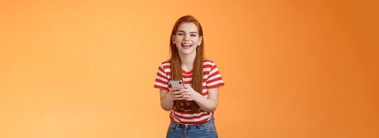 Carefree attractive woman red long hair laughing amused, having fun, chatting friend, hold smartphone, look camera sincere rejoice smile, texting joke, found funny meme internet, orange background photo