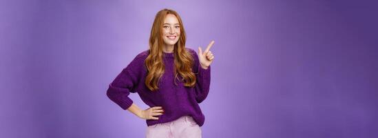 Hey click here. Portrait of friendly-looking charming and cute redhead female in purple sweater pointing at upper left corner with index finger and smiling, showing copy space, recommending, offering photo