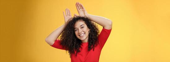 Close-up shot of charismatic playful and tender young kind woman with curly hair playing having fun showing bunny ears with hands on head tilting head joyfully fooling around and smiling at camera photo