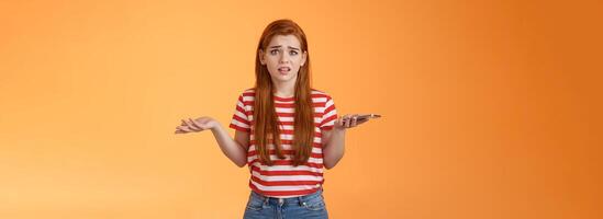 Complicated nervous redhead insecure girl receive strange message look perplaxed, unaware what do, spread hands sideways, hold smartphone, growning bothered worried, standing confused photo