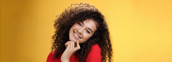 Close-up shot of charming flirty and silly curly-haired young woman making faces as trying get what wants, smiling tilting head on shoulder and touching lip standing cute over yellow background photo