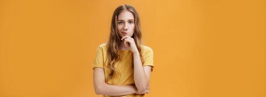 Hmm choices difficult. Portrait of focus serious-looking creative young female with wavy hair smirking holding hand on chin in thoughtful pose, making up decision, thinking over orange background photo