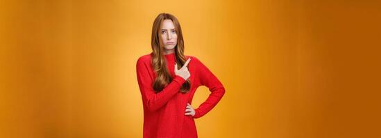Upset and disappointed young ginger girl in red sweater pointing at upper left corner or behind frowning looking gloomy and angry, standing jealous and moody against orange background photo
