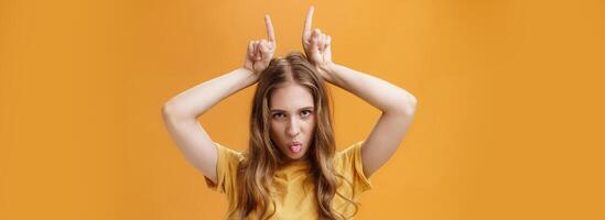 Daring and cool emotive attractive female student with wavy fair hair and little scars showing tongue rebellious looking from under forehead holding index fingers on head being stubborn photo