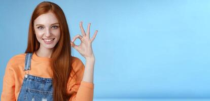 Sassy confident redhead girl got under control show okay ok excellent gesture cheeky look camera grinning assertive assuring everything awesome give permission approve good product, blue background photo