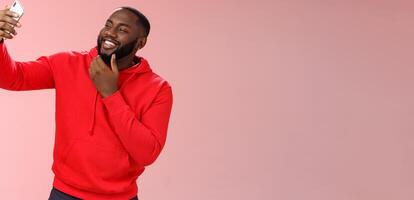 Cheeky cool african american confident bearded guy in red hoodie taking selfie update date app profile photo holding smartphone raised touching face sassy smiling look phone display flirty