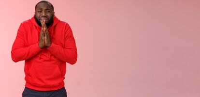 Miserable african american man beard in red hoodie stooping make supplicating upset grimace grying sobbing asking help say please hold hands pray begging mercy, apologizing need money photo