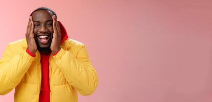 Happy charismatic playful african american bearded guy in yellow jacket red hoodie press palms cheeks smiling friendly laughing happily joking cannot believe receive awesome surprise, winning lottery photo