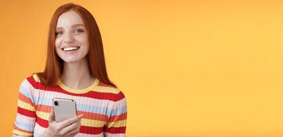Friendly enthusiastic young redhead girl blue eyes using smartphone turn camera answer smiling broadly telling who sent message standing delighted orange background messaging, texting boyfriend photo