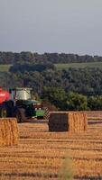 Haystacks on the field and tractor working on the natural green background. Dry straw pressed into square bales by agricultural tractor. Tractor pressing grass for livestock in summer. Vertical video