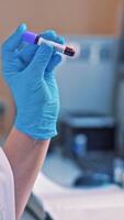 Laboratory worker holds a vial and mixes it by hand in hospital. Technician woman works with blood samples while standing close to the automated machine in clinic. Close-up. Vertical video
