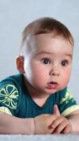 Funny little kid lying on his belly on the floor looks into camera his eyes wide open. Lovely child looking sideways and rubbing tiny fists. Vertical video