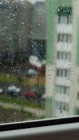 Rain drops on window glass in viewing cityscape. Drops of rain on a window pane with buildings background. Raining scenery on window. Vertical video
