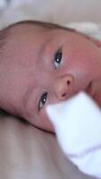 Newborn kid in white costume lying on the bed and looking with his eyes wide-open. Baby puts his hand in front of his mouth. Close-up. Vertical video