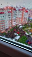 View through the window on raining weather. Drops of rain on a window pane on the city buildings background. Rain droplets flowing on a glass. Vertical video