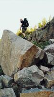 A traveler in shorts and with a backpack climbs onto a huge stone and sits on it amid a quarry with a lake in the center. Vertical video
