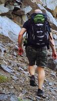 A traveler with a backpack on his shoulders rises to the top of the mountain with stones in the afternoon in warm weather. Tourism. Vertical video