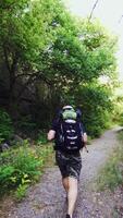 A man with a backpack on his shoulders is climbing up the path against the background of green trees in the summer on a warm day. Tourist in a hike. Vertical video