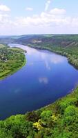 A wide blue river flows between rocks with trees and countryside with long fields and a private sector. Aerial view. Vertical video