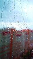 Raindrops on window in the city. Rainy weather with a sun on the sky. Water drops flowing on the glass window on the background of high-rise buildings. Vertical video