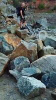 A tourist in shorts and a backpack is standing on a huge stone in the middle of a quarry and looking into the distance to the surrounding landscape. Camera motion up. Aerial view. Vertical video