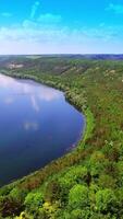 View of the countryside with houses, fields and trees which are surrounded by wide river on the background of high rocks. Aerial view. Vertical video