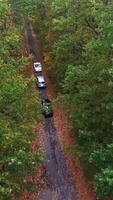 Aerial view of three cars moving along the narrow road between colored trees in the forest. Road with cars surrounded by trees in autumn. Motion bottom up Vertical video