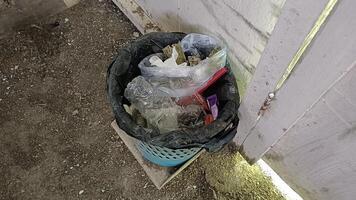 A black trash bag overflowing with garbage, including plastic bottles, plastic bags, and other assorted trash, sits in a blue trash can against a white wooden fence. photo
